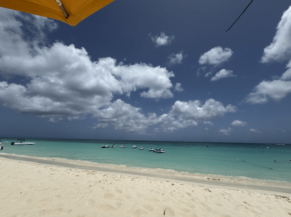A sandy beach with turquoise water and a row of jet skis near the shore under a partly cloudy blue sky. The edge of a yellow umbrella appears in the upper left—just another day for my honest travel blog, It’s How I Travel.